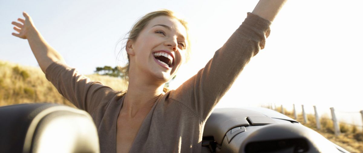 Young Woman Sits in the Back of a Convertible, Her Arms in the Air, Laughing With Joy