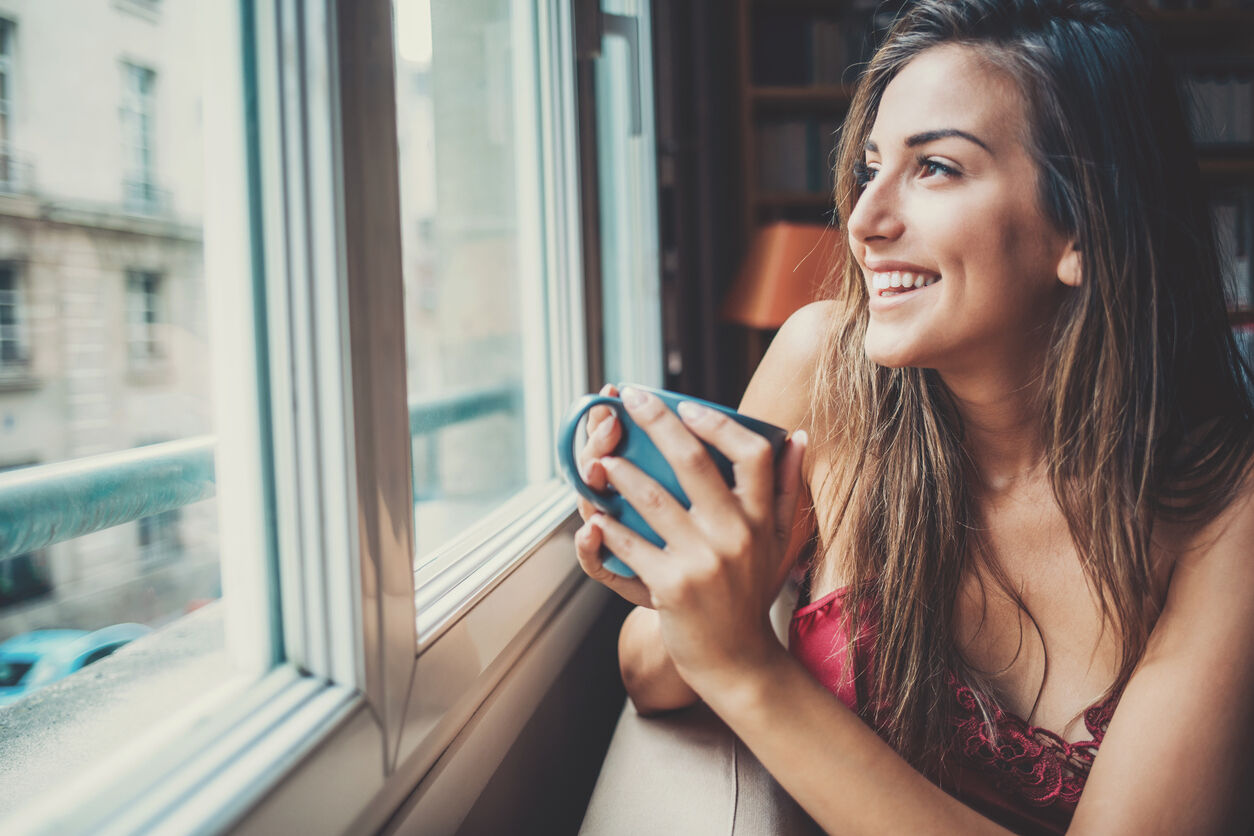 Beautiful young woman holding a cup of coffee looking through a house window. Shot made during Istockalypse Paris 2016 event.