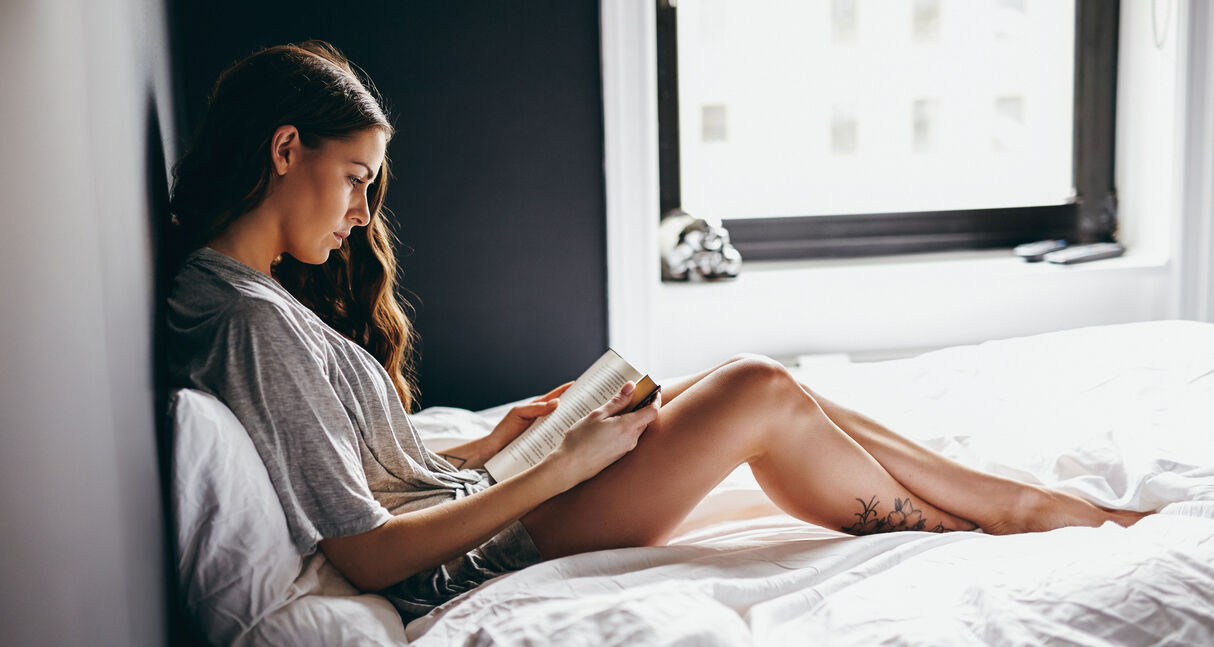 Side view of beautiful young woman reading book on bed at home. Female model in nightwear sitting on bed read a novel.