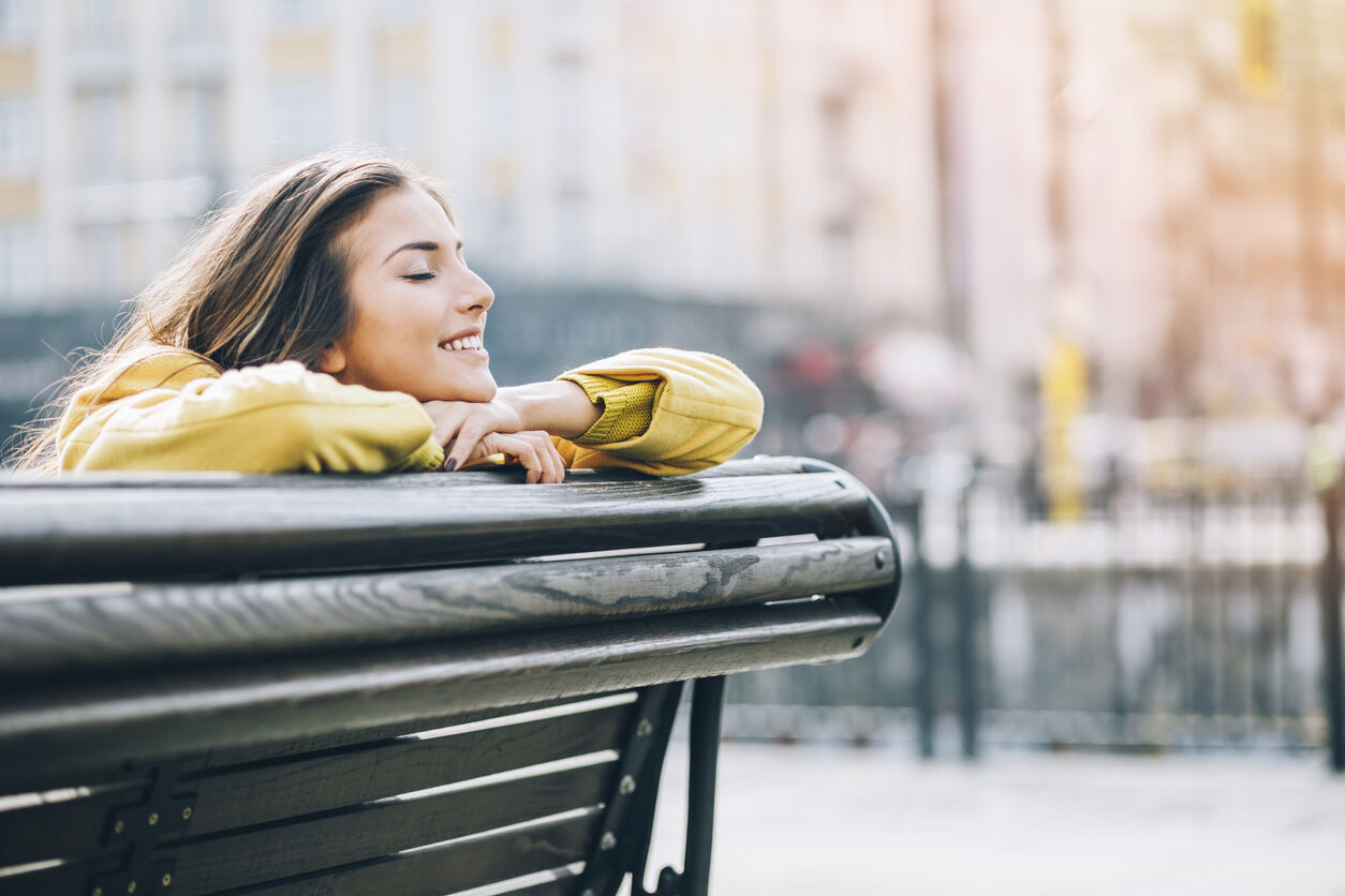 Young woman smiling with eyes closed, sitting on a bench outdoors under the sunlight, with copy space