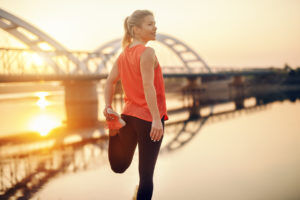 Cute Caucasian blonde woman in sportswear stretching leg by the river. It's hard to beat a person who never gives up.
