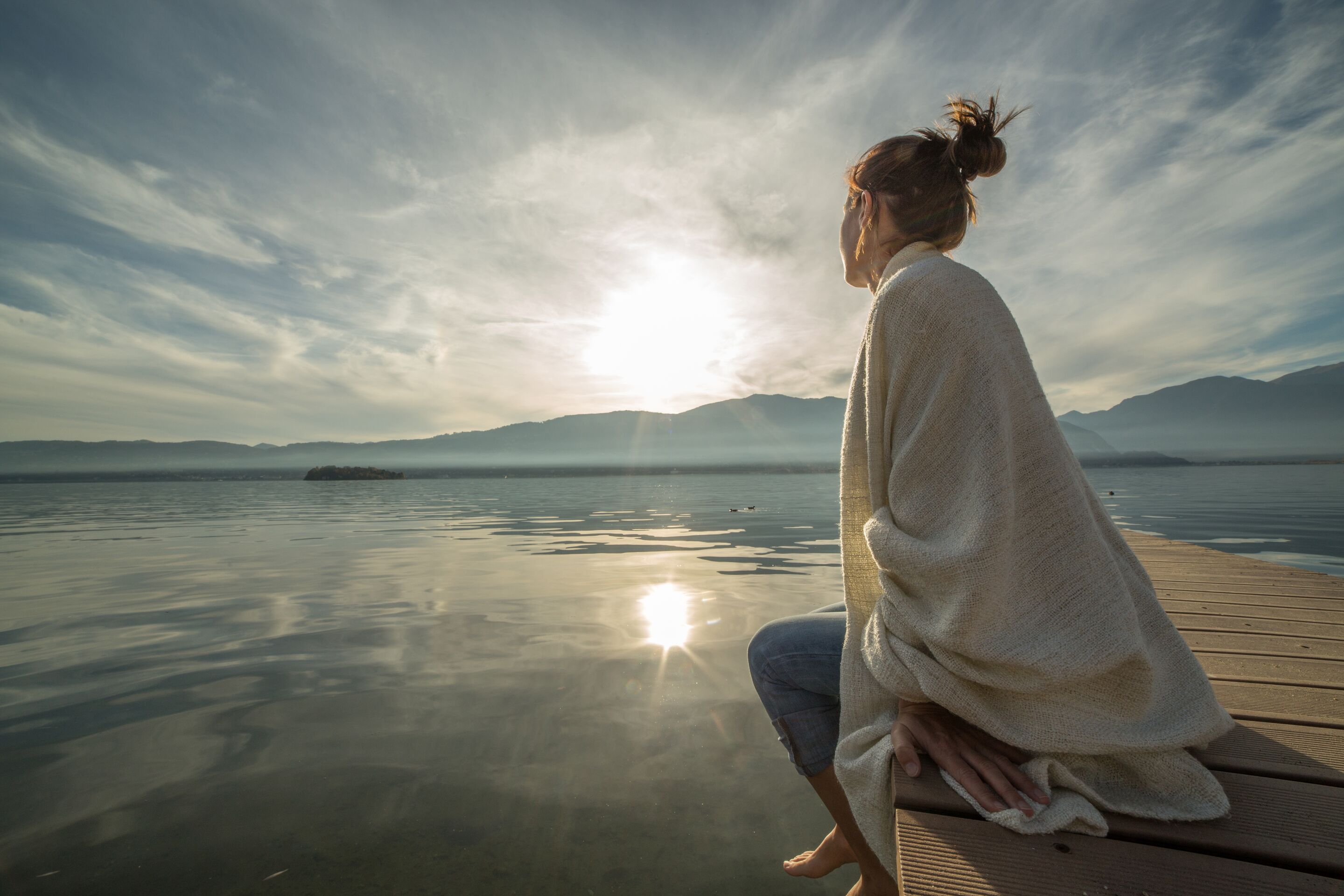 Young woman relaxes on lake pier with blanket, watches sunset