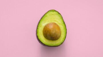 Een halve avocado met bruine pit in het midden tegen een roze achtergrond.