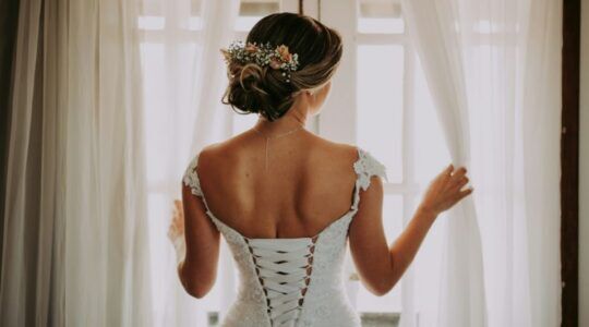 Een bruid staat met haar rug naar de camera toe voor witte gordijnen.