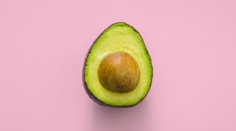 Een halve avocado met bruine pit in het midden tegen een roze achtergrond.